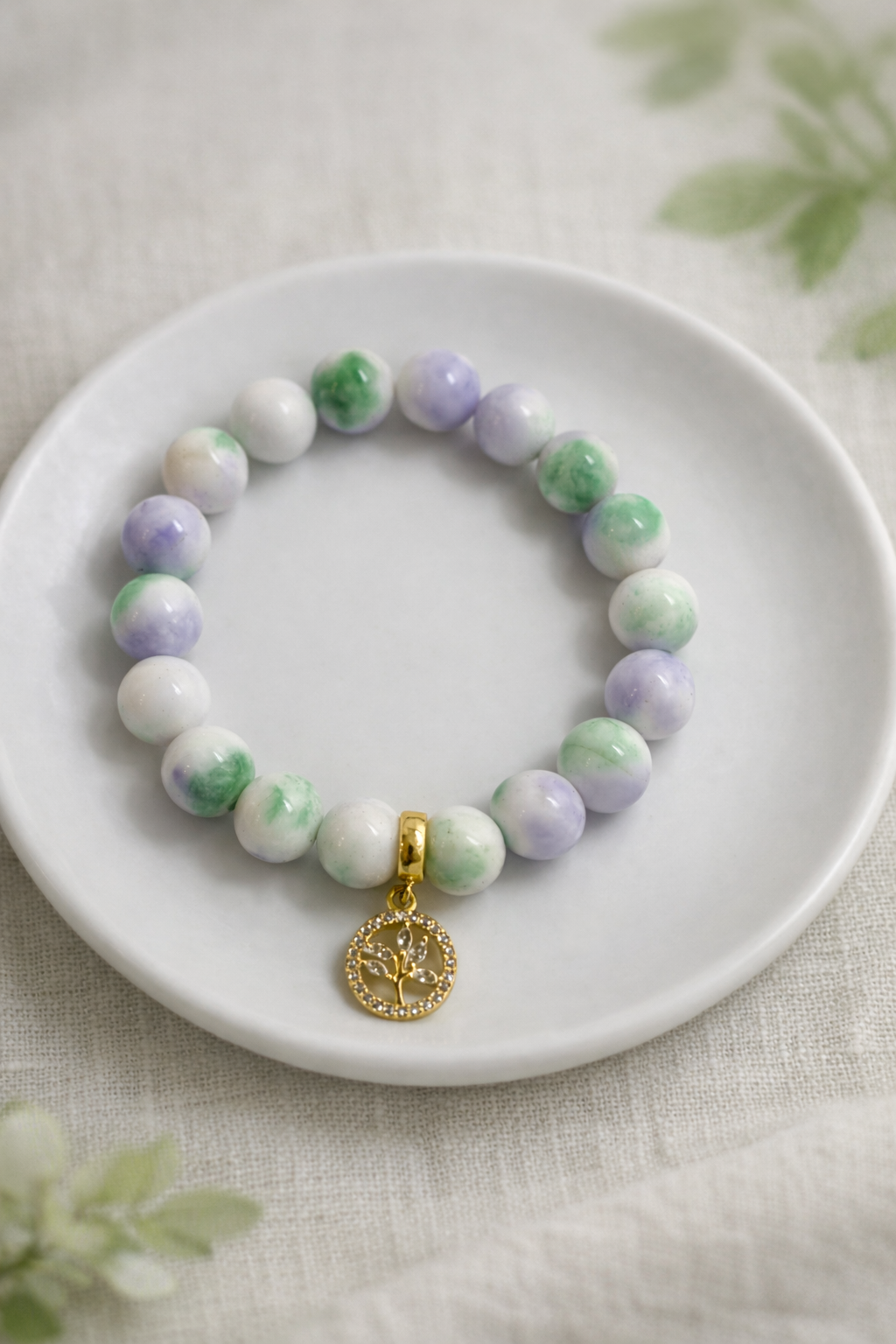 Minimālistiska dabīgo akmeņu aproce ar zaļiem, baltiem un gaiši violetiem pērlēm, novietota uz balta keramikas šķīvīša. Aprocei pievienots zelta “Dzīvības koka” kulons ar smalkiem kristāliem. Fons – gaišs lina audums ar maigi izpludinātiem zaļiem augu elementiem, maigs dabīgais apgaismojums un sekls asuma dziļums.