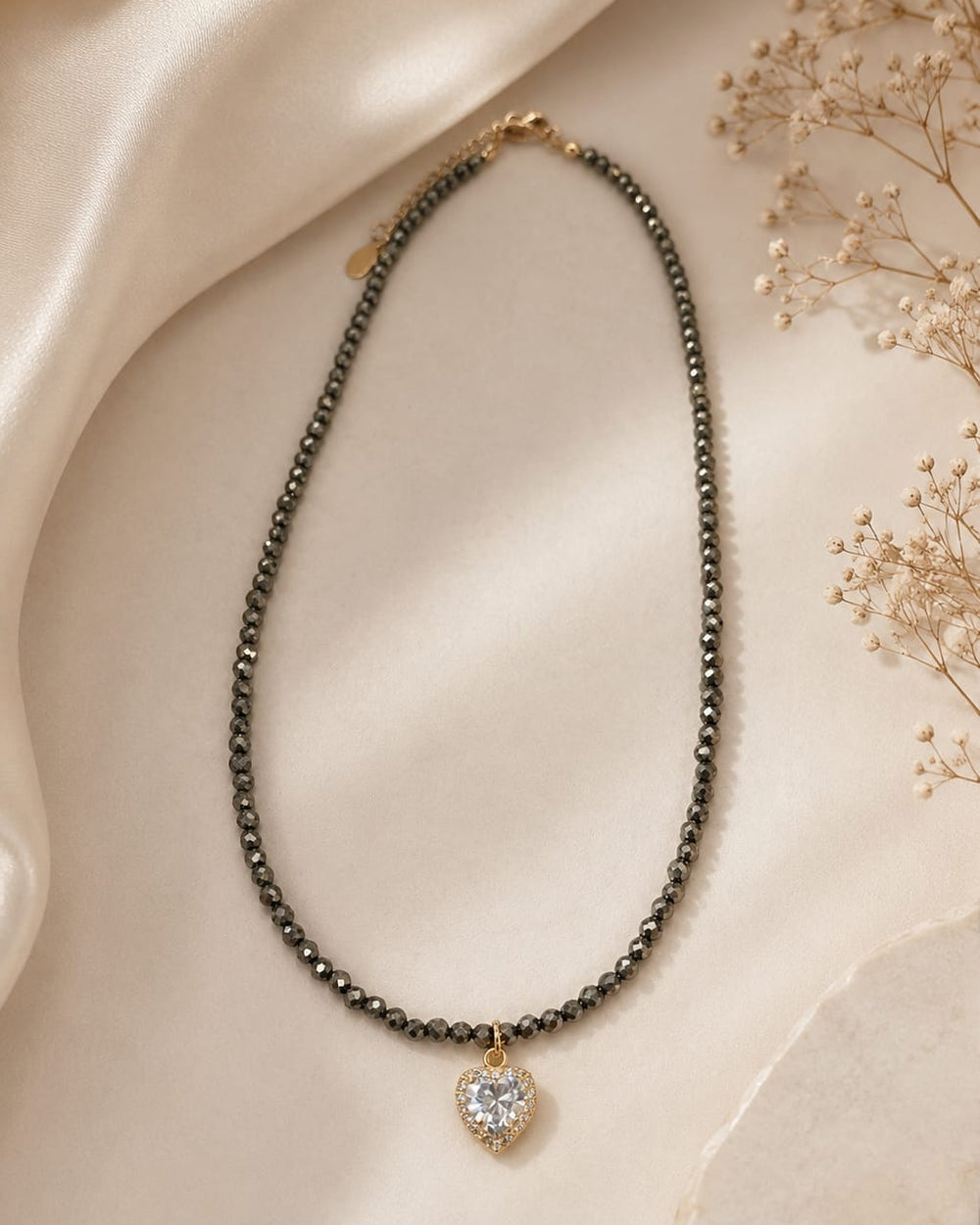 Eleganta 3 mm pirīta kaklarota ar mirdzošu sirds formas piekariņu un regulējamu zeltītu aizdari, novietota uz gaiša, minimālistiska fona. Dabīgā gaisma izceļ pirīta akmeņu spīdumu un sirds piekariņa mirdzumu, radot romantisku un greznu luxury editorial noskaņu.