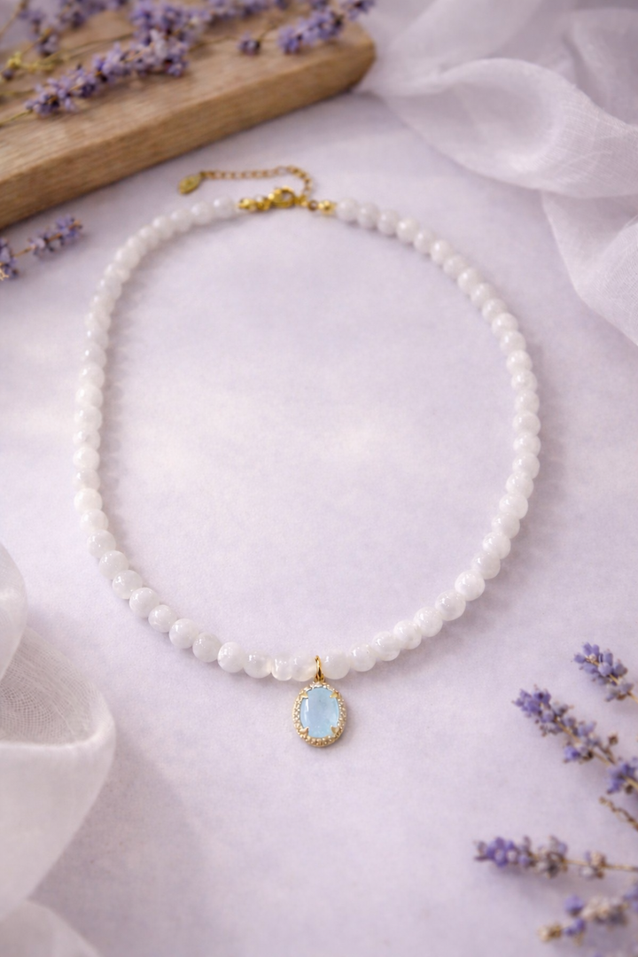 8 mm mēnessakmens bumbiņu kaklarota ar ovālu gaiši zilu akvamarīna kulonu apzeltītā ietvarā, romantiskā kompozīcijā uz maigi violeta fona ar lavandas ziediem.