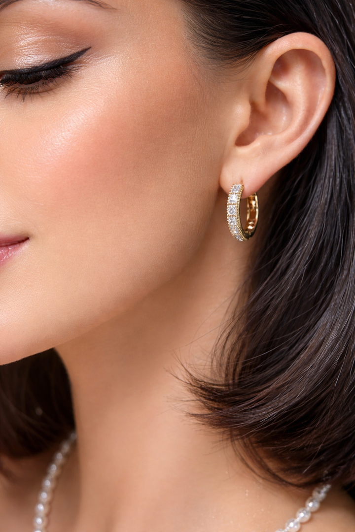 Koķets, pavasarīgs modeles tuvplāns ar maigu smaidu, kur skaidri redzama seja un zelta auskari ar mirdzošiem akmentiņiem; fonā maigas margrietiņas un viegla, svaiga noskaņa.