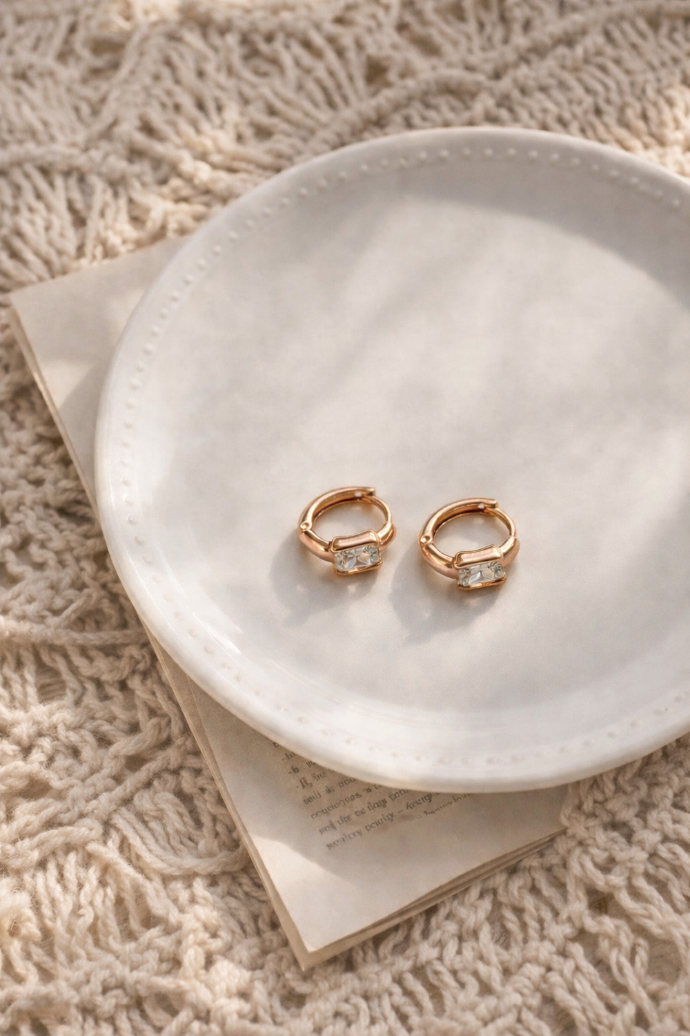 Gaišā rozā zelta mini riņķa auskari ar taisnstūra dzidru kristālu uz baltas keramikas šķīvja ar punktotu maliņu; zem šķīvja redzama grāmatas lapa, bēšs makramē fons.