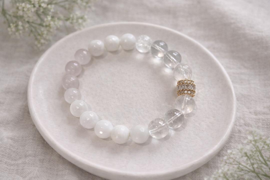 Minimālistiska produktu fotogrāfija ar rokassprādzi no baltiem un caurspīdīgiem dabīgā akmens pērlēm ar zelta krāsas starp elementu ar kristāliem. Aproce novietota uz gaiša keramikas šķīvīša, fonā dabīgs lina audums un maigi izpludināti augu elementi. Apgaismojums maigs, dabīgs, radot elegantu un harmonisku rotas prezentāciju. ✨