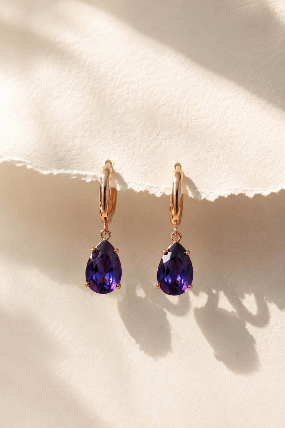 Apzeltīti auskari ar angļu aizdari un violetiem lāsītes formas kristāliem uz gaiša teksturēta papīra fona. Eleganti sieviešu auskari minimālistiskā luxury stilā ar siltu dabīgo apgaismojumu.