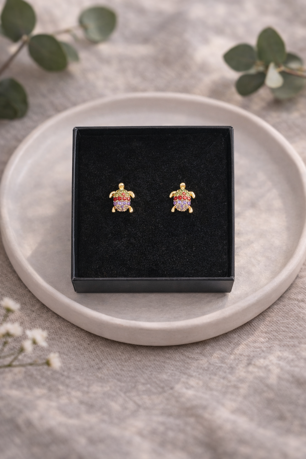 Krāsaini bruņurupuča formas auskari ar kristāliem, ievietoti melnā rotaslietu kastītē uz gaiša keramikas šķīvīša, minimālistiska rotaslietu produktu fotogrāfija ar dabīgu lina fonu un maigu dienasgaismu.