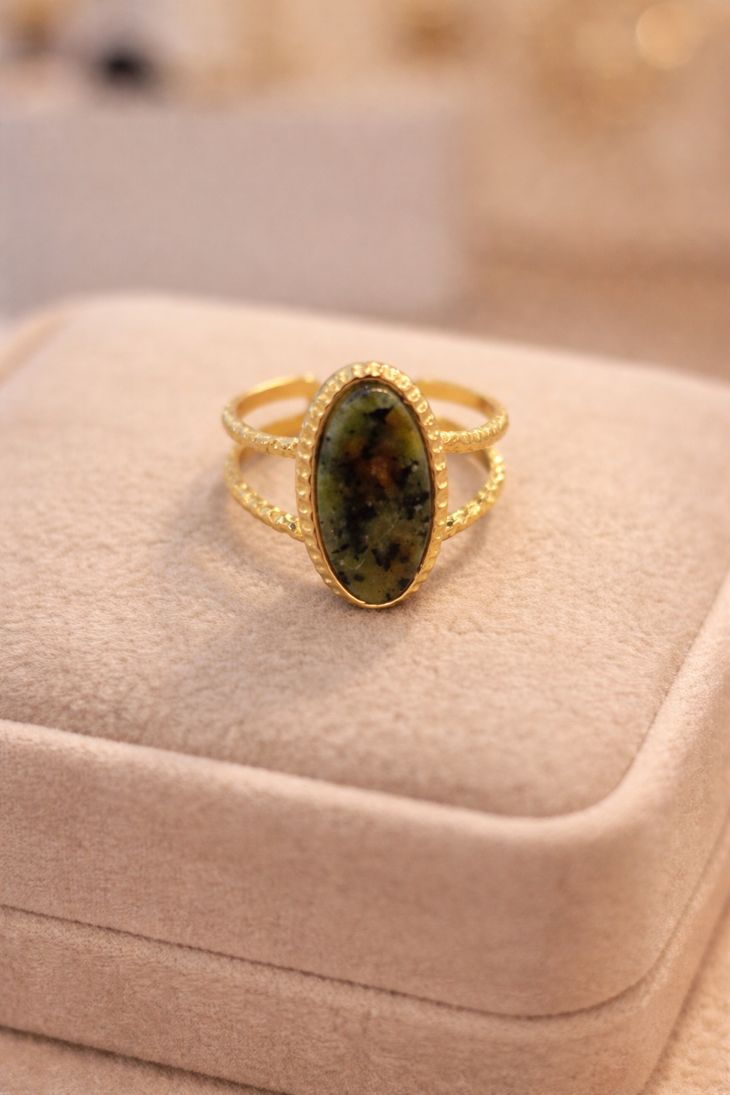 Zelta toņa gredzens ar ovālu zaļgani brūnu dabīgā akmens ielaidumu un dubultu stīpiņas dizainu, novietots uz gaišas samta juvelierizstrādājumu kastītes siltā, maigi izkliedētā apgaismojumā.