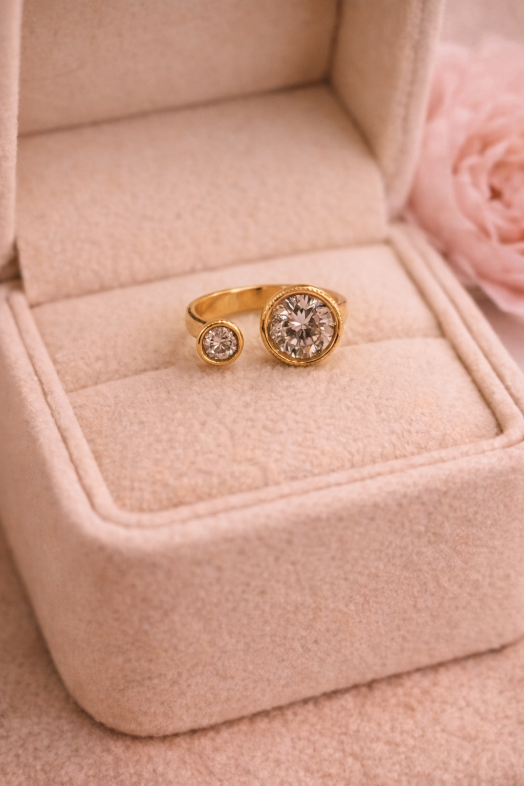 Zelta krāsas atvērta tipa gredzens ar diviem apaļiem dzidriem kristāliem – vienu lielāku un vienu mazāku – novietots uz gaišas samta rotaslietu kastītes siltā, maigi izpludinātā apgaismojumā.