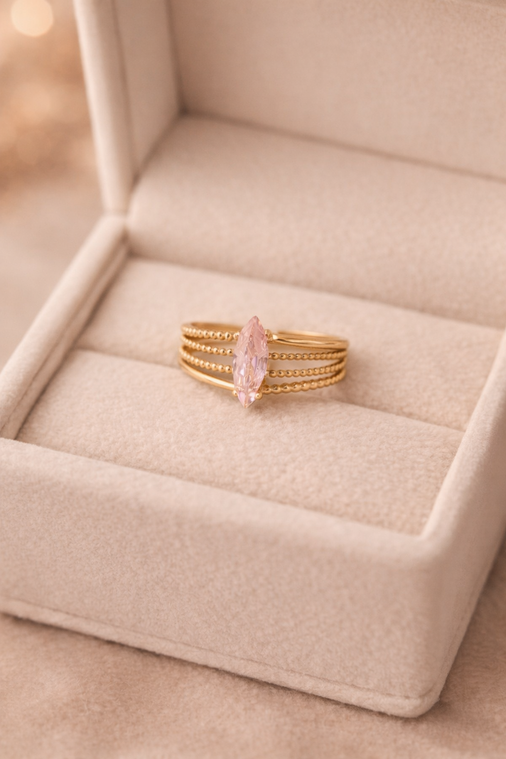 Zelta krāsas gredzens ar vairākām smalki teksturētām joslām un centrā novietotu rozā, marķīzes (iegareni lāsītes) formas kristālu, izvietots uz gaišas samta juvelierizstrādājumu kastītes siltā apgaismojumā.