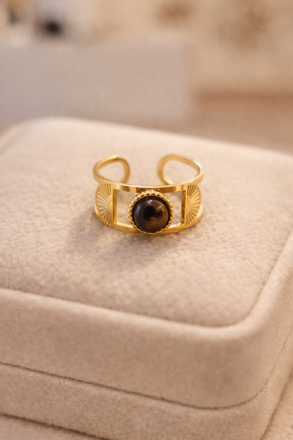 Atvērta dizaina zelta gredzens ar centrā iestrādātu brūnu, apaļu akmeni un dekoratīvām reljefa detaļām sānos, novietots uz gaišas samta juvelierizstrādājumu kastītes siltā, maigā apgaismojumā.