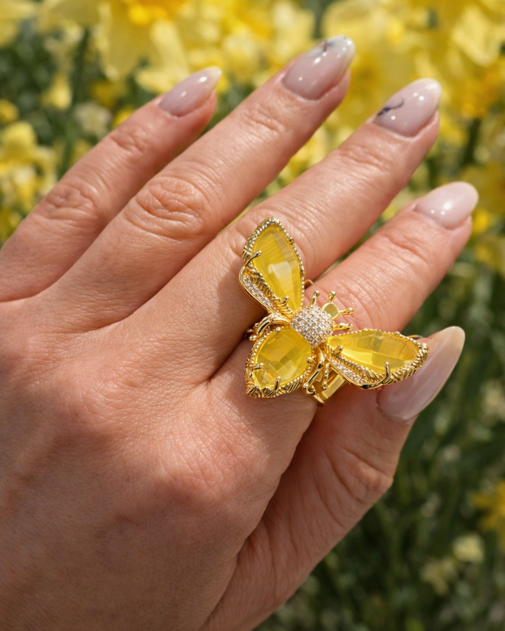 Sievietes roka ar lielu tauriņa formas gredzenu ar dzelteniem slīpētiem akmeņiem un zelta krāsas ietvaru uz pavasara dzeltenu ziedu fona.