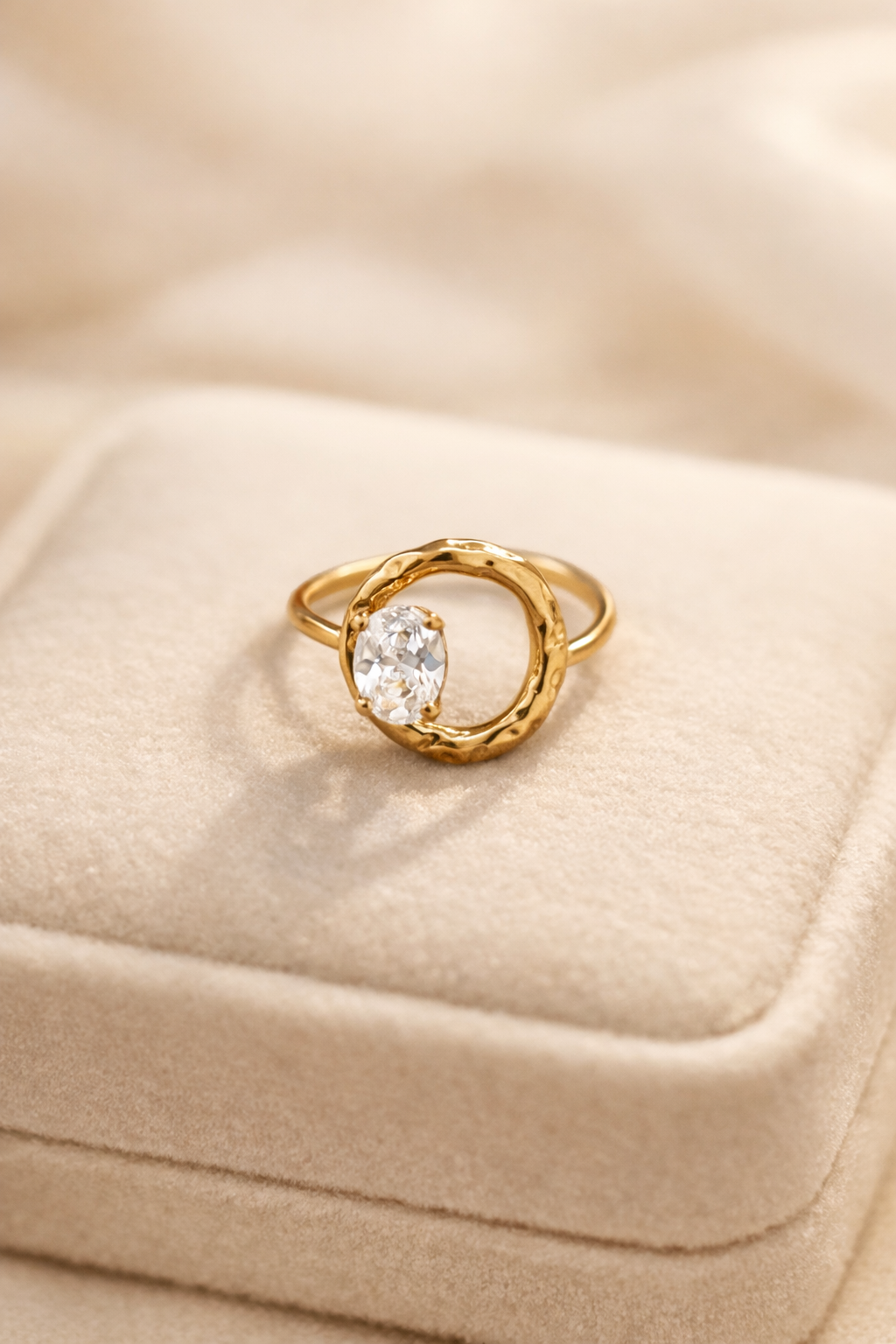 Zelta gredzens ar apaļu, teksturētu ietvaru un centrā iestrādātu dzidru ovālu kristālu, novietots uz gaišas samta juvelierizstrādājumu kastītes, maigs silts apgaismojums un izpludināts fons.