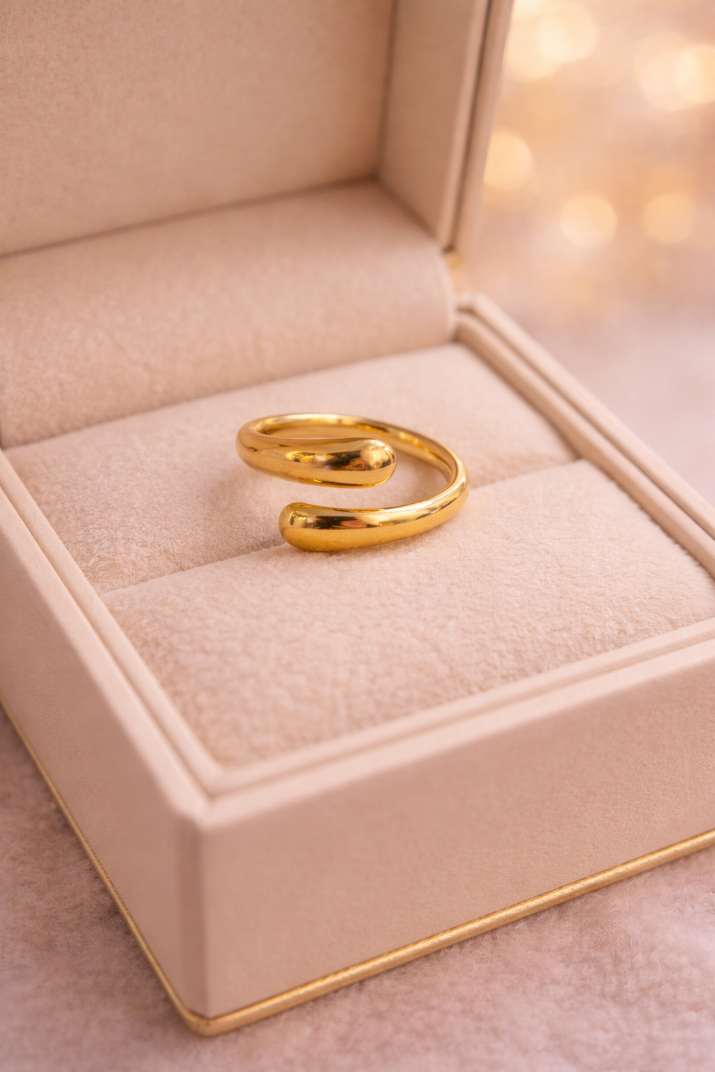 Zelta krāsas atvērta dizaina gredzens ar gludu, noapaļotu formu, novietots uz gaišas samta juvelierizstrādājumu kastītes, silts un maigs apgaismojums ar izpludinātu fonu.
