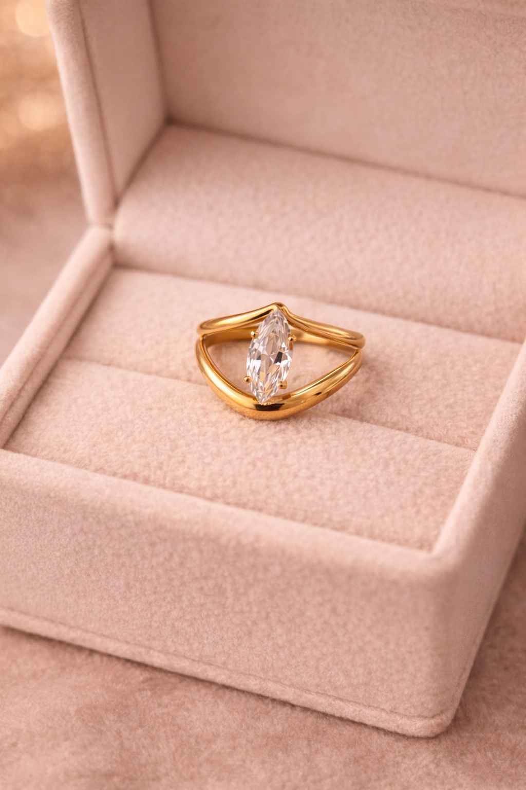 Zeltains gredzens ar marķīzes (iegarenas lāsītes) formas caurspīdīgu kristālu, novietots uz gaišas samta rotu kastītes siltā, maigi izpludinātā apgaismojumā.