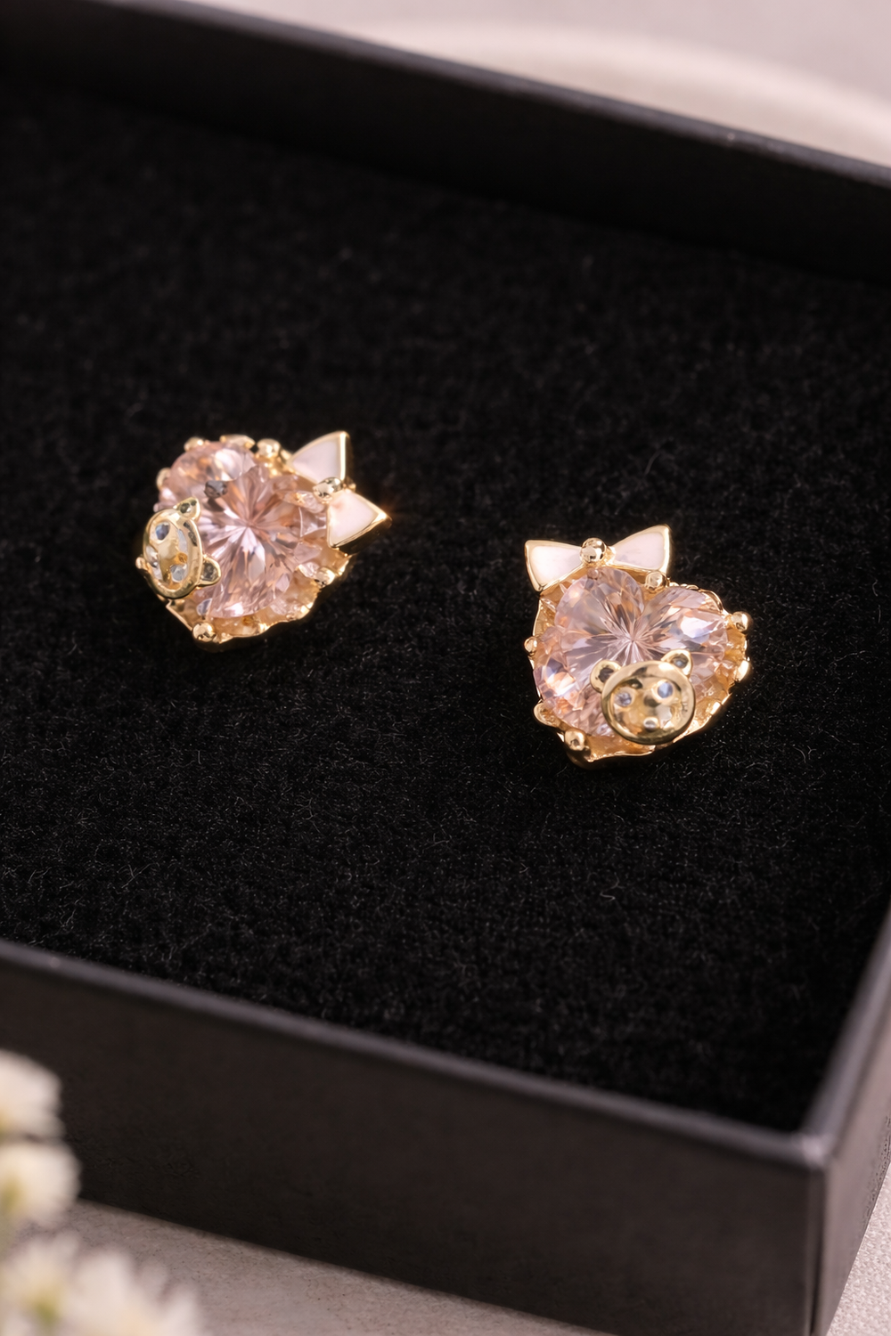 Tuvplāna fotogrāfija ar zelta krāsas bērnu auskariem ar rozā kristāliem melnā rotaslietu kastītē, redzamas smalkas detaļas un kristāla mirdzums uz maiga, izpludināta lina fona.