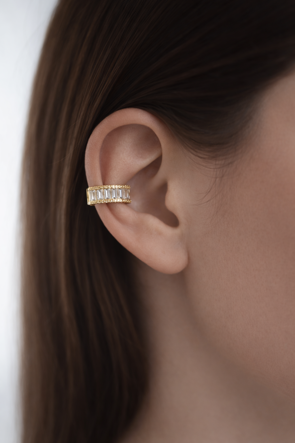 Elegants zelta ear cuff tipa auskars ar taisnstūrveida kristāliem, uzlikts uz sievietes auss ārējās malas. Mati gludi, brūni un aizlikti aiz auss, fons gaišs un maigi izpludināts, fokuss uz rotas detaļām.