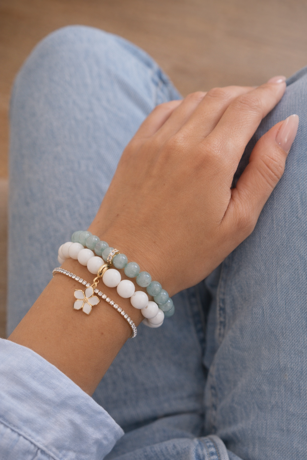 Sievietes roka ar gaiši rozā manikīru, uz kuras redzamas trīs rokassprādzes – gaiši zilu un baltu akmeņu aproces ar zieda piekariņu un smalka tenisa aproce. Roka novietota uz ceļa, fonā gaiši zili džinsi un maigi izpludināts silts interjers.