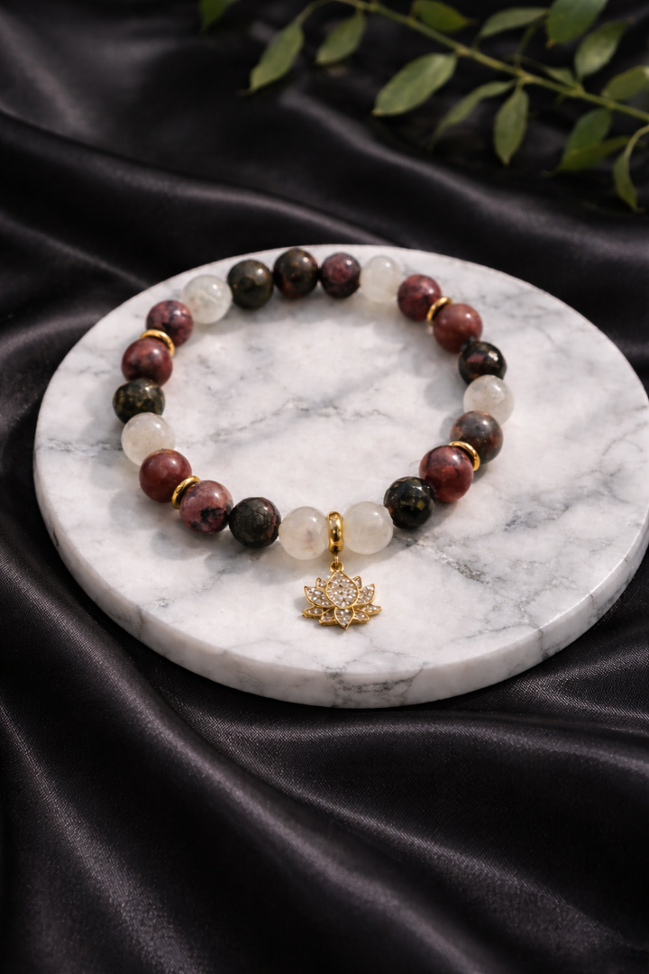 Roku darbs dabīgo akmeņu aproce ar mēnessakmens, pirīta un rodonīta pērlēm un zelta lotosa piekariņu, novietota uz balta marmora paliktņa ar melna zīda fonu un zaļu augu zaru fonā.