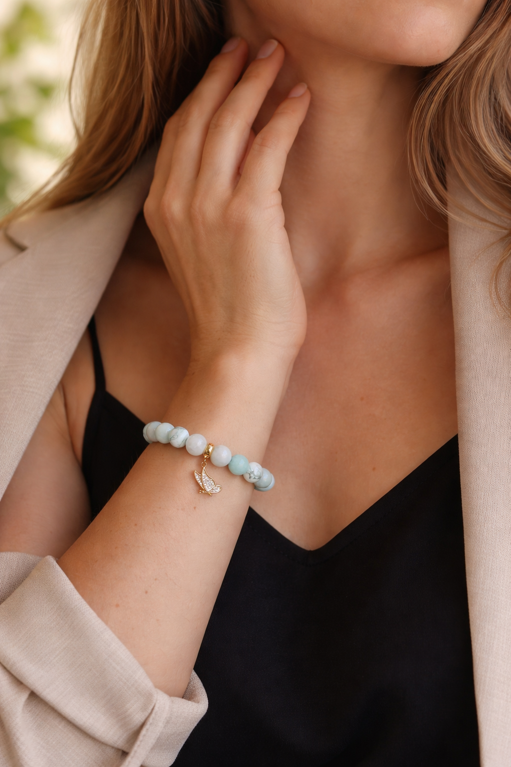 Sieviete melnā topā un gaišā bēšā žaketē ar roku maigi pieskaras kaklam. Uz plaukstas – dabīgo akmeņu rokassprādze ar hovlīta, mēnessakmens un amazonīta pērlēm un zelta tauriņa piekariņu. Silts, dabīgs apgaismojums, izpludināts zaļš fons.