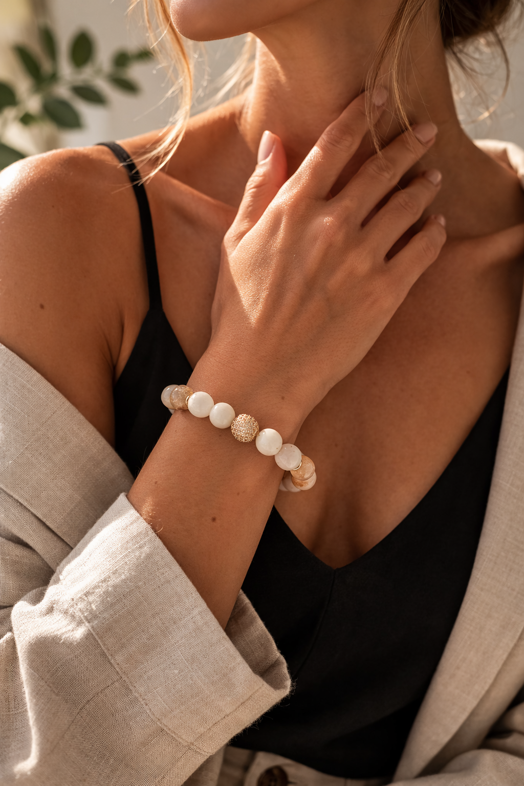 Sievietes rokas tuvplāns pie kakla siltā dabīgā apgaismojumā. Uz plaukstas balta un bēša dabīgo akmeņu aproce ar zelta akcenta bumbiņu. Sieviete valkā melnu topu ar plānām lencītēm un gaišu bēšu žaketi, fons maigi izpludināts ar zaļu augu elementu.