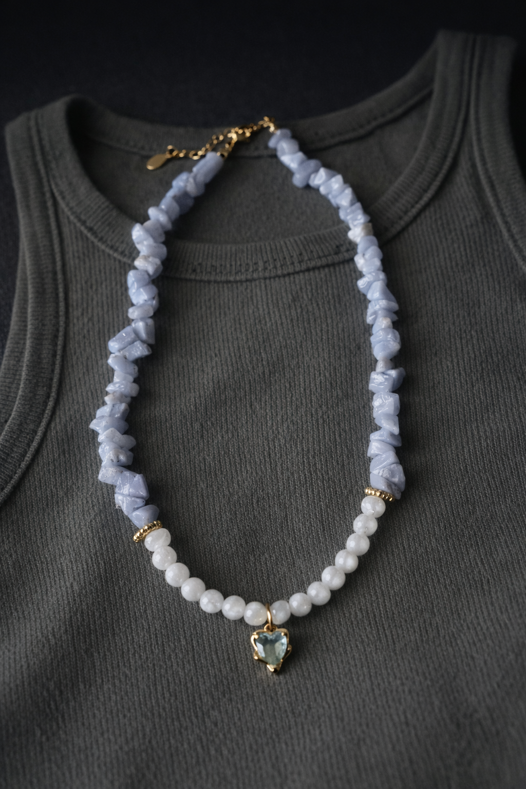 Kaklarota ar gaiši ziliem akmens čipsiem un baltām pērlītēm, ar zeltītu sirds formas kulonu, izkārtota uz tumši pelēka bezpiedurkņu topa ar izteiktu auduma tekstūru; mīksts, kontrastains apgaismojums un tumšs, minimālistisks fons.