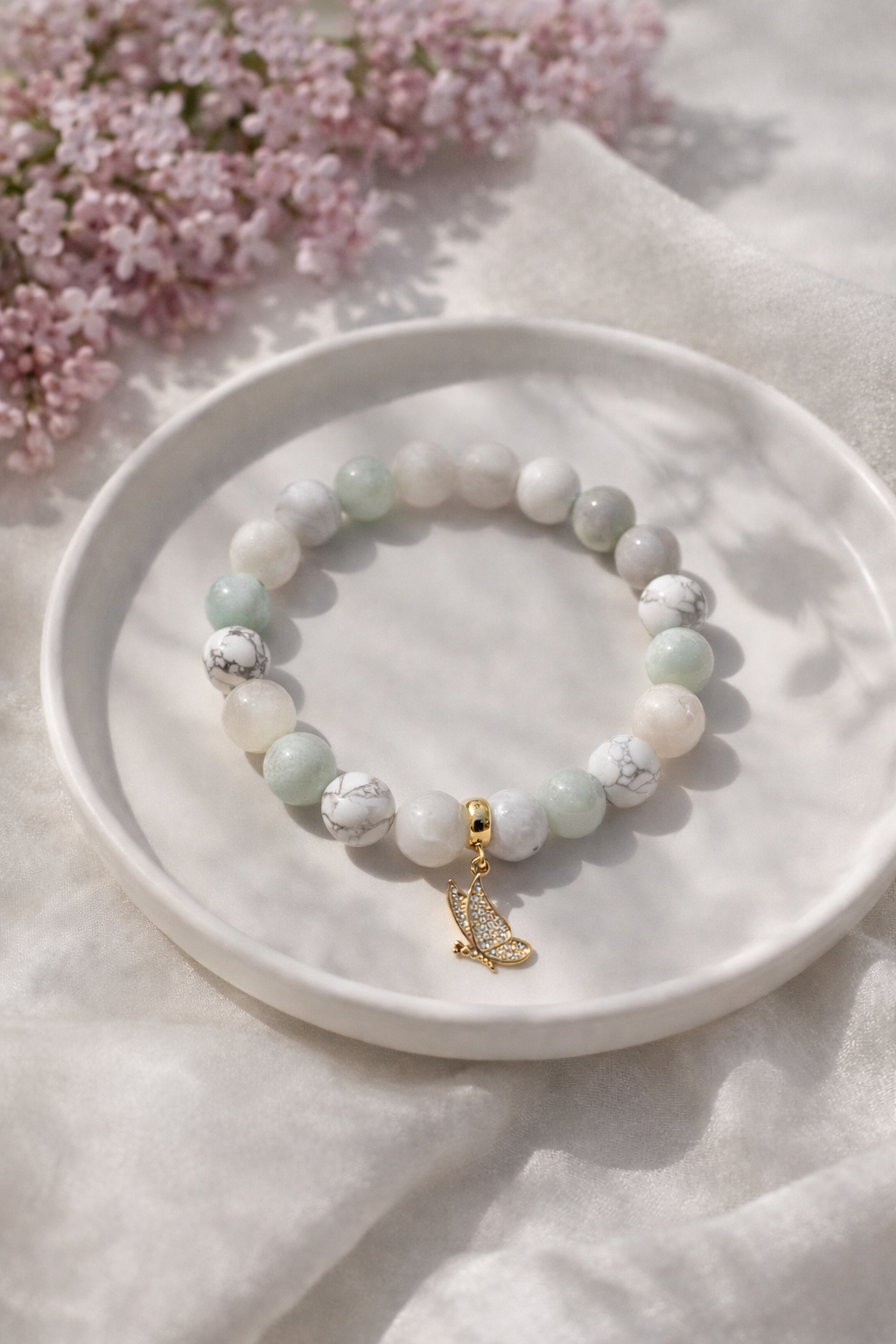 Dabīgo akmeņu rokassprādze ar hovlīta, mēnessakmens un amazonīta pērlēm un zelta tauriņa piekariņu, novietota uz baltas keramikas šķīvīša. Fons gaišs ar maigi rozā ceriņu ziediem un dabīgu saules ēnu efektu.