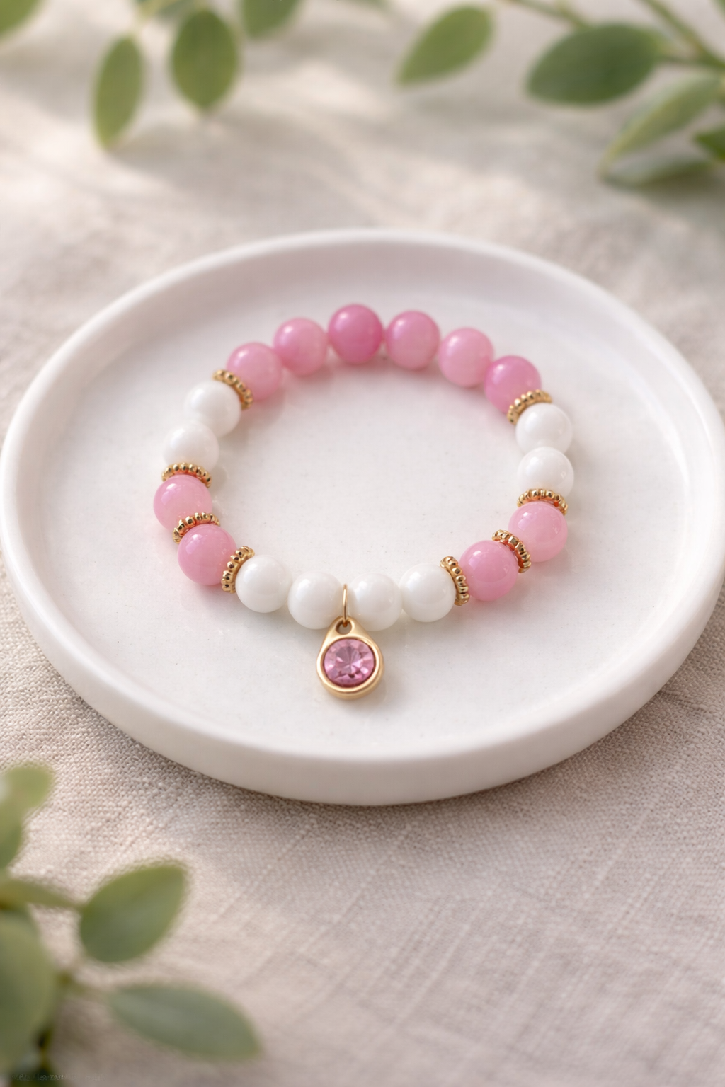 Rozā un baltudabīgā akmens bumbiņu rokassprādze ar zelta krāsas akcentiem un piekariņu ar rozā kristālu, novietota uz balta keramikas šķīvīša uz gaiša lina auduma fona ar izpludinātiem zaļiem augu elementiem, maiga dabīgā apgaismojumā.