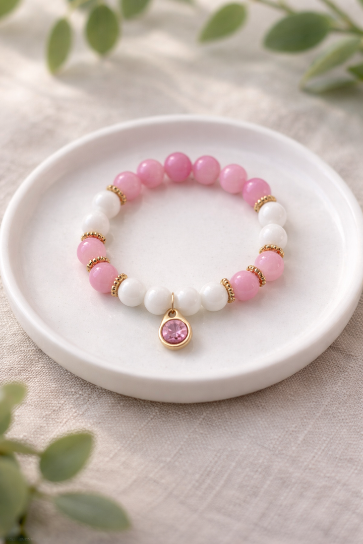 Rozā un baltudabīgā akmens bumbiņu rokassprādze ar zelta krāsas akcentiem un piekariņu ar rozā kristālu, novietota uz balta keramikas šķīvīša uz gaiša lina auduma fona ar izpludinātiem zaļiem augu elementiem, maiga dabīgā apgaismojumā.