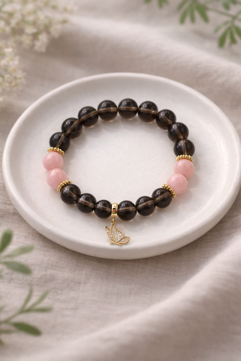 Minimālistiska rokassprādze no dūmakaina kvarca un rozā akmens pērlēm ar zelta tauriņa piekariņu, novietota uz balta keramikas šķīvīša uz gaiša lina auduma ar izpludinātu zaļu augu fonā.