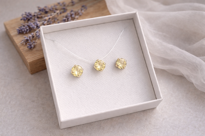 Minimālistiska rotu komplekta produktu fotogrāfija baltā kastītē – zeltītā ietvarā iestrādāti dzeltenas krāsas kristāli.Komplektā nagliņu auskari un kaklarota uz caurspīdīgas auklas. Fons gaišs ar koka dēlīti, žāvētu lavandu un viegli plīvojošu caurspīdīgu audumu, maigs dabīgais apgaismojums un sekls asuma dziļums.