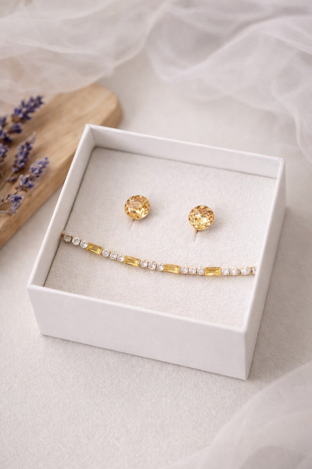 Balta rotaslietu kastīte ar baltu samta iekšpusi, kurā izvietoti apaļi zeltaini mirdzoši auskari un zelta tenisa stila rokassprādze ar dzelteniem un dzidriem akmentiņiem; fonā gaiša matēta virsma, koka dēlītis ar žāvētu lavandu un viegli izpludināts caurspīdīgs audums.