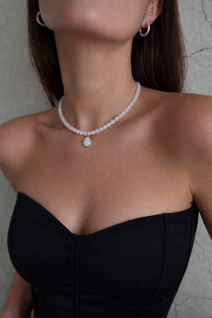 Mēnessakmens 8 mm bumbiņu kaklarota ar nelielu gaiši zilu akvamarīna kulonu apzeltītā ietvarā, redakcionālā tuvplānā uz sievietes kakla un pleciem, valkāta ar melnu korsetes tipa topu uz neitrāla pelēka fona.