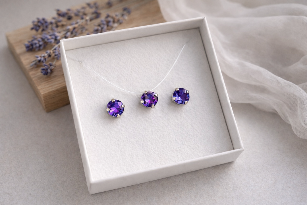 Minimālistiska rotu komplekta produktu fotogrāfija baltā kastītē – sudraba ietvarā iestrādāti zili-violeti kristāli. Komplektā nagliņu auskari un kaklarota uz caurspīdīgas auklas. Fons gaišs, ar koka dēlīti, žāvētu lavandu un viegli izpludinātu audumu, maigs dabīgais apgaismojums.