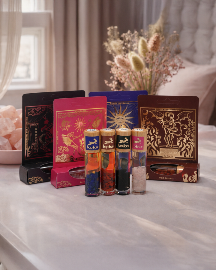 Estētiska produktu fotogrāfija ar četriem ēterisko eļļu rullīšiem ar dabīgiem kristāliem, kas novietoti priekšplānā. Aizmugurē krāsainas, dekoratīvas iepakojuma kastītes ar zodiaka un simboliskiem motīviem. Silta, dabiska gaisma un maigi izpludināts fons ar ziediem rada mistisku, harmonisku un nedaudz ezotērisku atmosfēru.