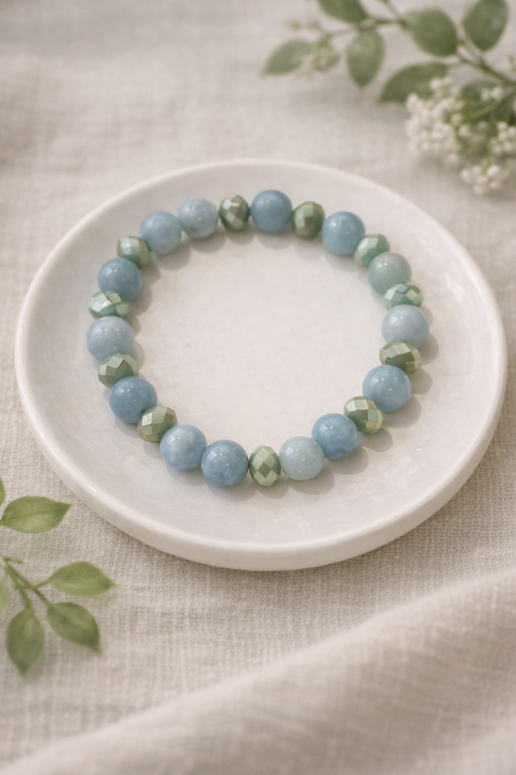 Gaiši zilas un maigi zaļas pērlīšu rokassprādze, novietota uz balta keramikas šķīvīša uz gaiša lina auduma, fonā maigi izpludināti zaļi augu zari un balti ziedi.