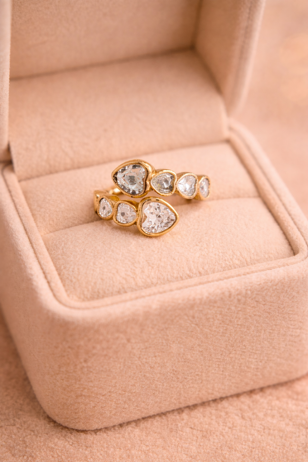 Zelta krāsas divrindu gredzens ar vairākiem dažāda izmēra caurspīdīgiem sirds formas kristāliem, novietots uz gaišas samta rotaslietu kastītes.