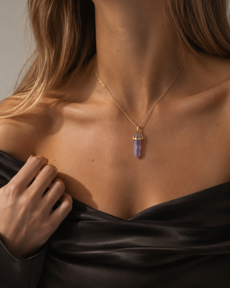 Sieviete melnā satīna off-shoulder topā valkā apzeltītu kaklarotu ar violeta lepidolīta kristāla kulonu. Silta dabīgā gaisma izceļ kaklarotas zelta mirdzumu un akmens maigo violeto toni.