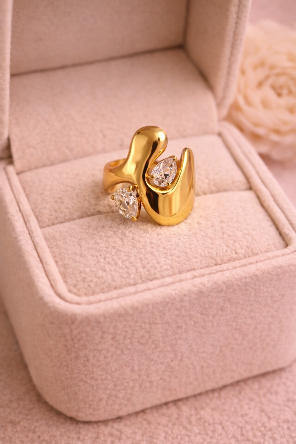 Zelta krāsas gredzens ar plūstošu dizainu un diviem dzidriem bumbiera formas kristāliem, novietots gaišā samta rotaslietu kastītē siltā apgaismojumā.