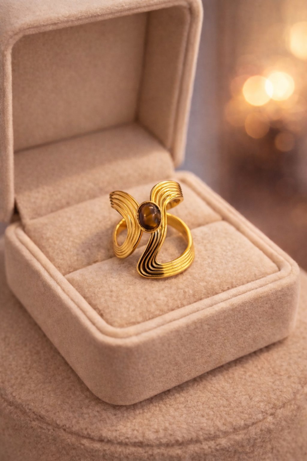 Zelta krāsas regulējams gredzens ar plūstošu, viļņveida dizainu un ovālu tīģeracs akmeni centrā, novietots gaišā samta rotaslietu kastītē siltā apgaismojumā.