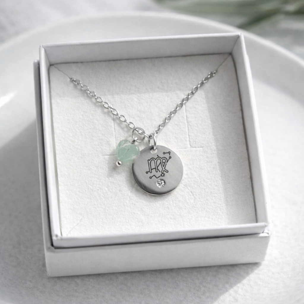 Sudraba kaklarota ar gravētu apaļu kulonu un mazu zaļganu akmens piekariņu, ievietota baltā rotaslietu kastītē uz balta keramikas šķīvja, ar maigu dabīgo apgaismojumu un izpludinātu augu fonā.
