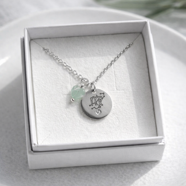 Sudraba kaklarota ar gravētu apaļu kulonu un mazu zaļganu akmens piekariņu, ievietota baltā rotaslietu kastītē uz balta keramikas šķīvja, ar maigu dabīgo apgaismojumu un izpludinātu augu fonā.
