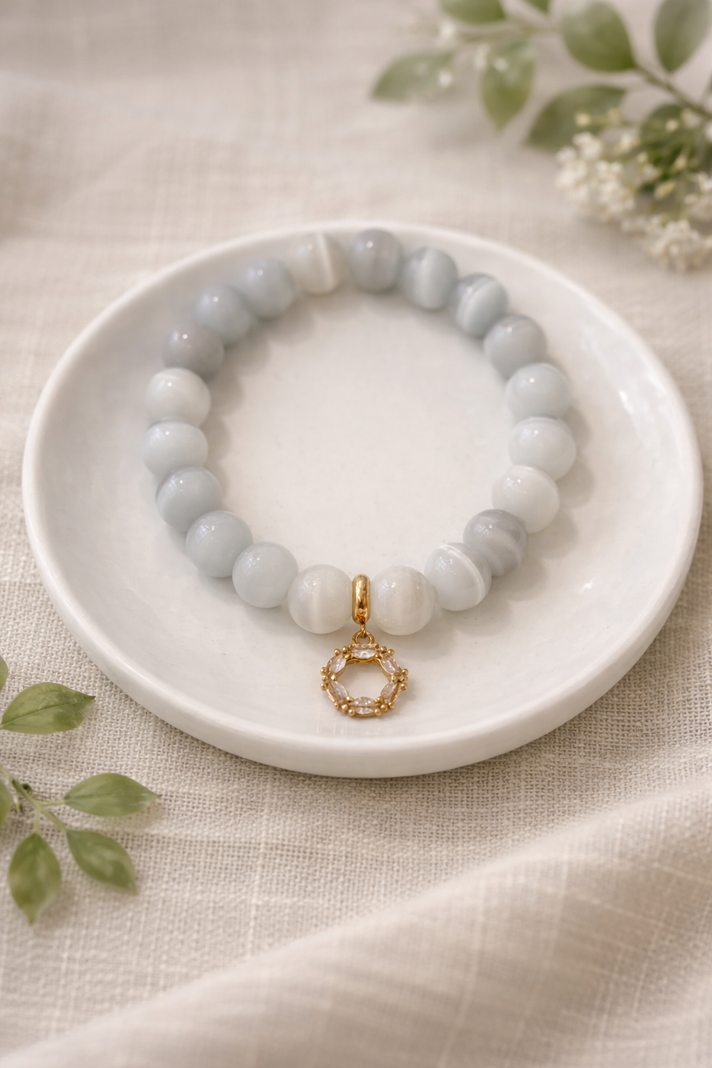 Balta un gaiši pelēka ahāta pērlīšu rokassprādze ar zelta krāsas dekoratīvu piekariņu, novietota uz balta keramikas šķīvīša uz gaiša lina auduma, fonā maigi izpludināti zaļi augi un balti ziedi.