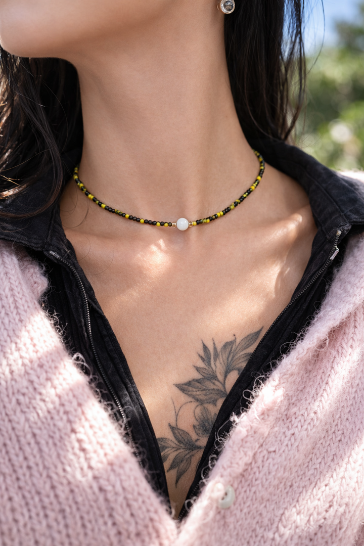 Tuvplāna lifestyle fotogrāfija ārā saulainā dienā ar sievietes kakla un atslēgas kaulu līniju fokusā. Ap kaklu – melni un dzelteni pērlīšu čokeris ar centrālo baltu mēnessakmens bumbiņu. Maigi rozā kardigans un melns tops ierāmē redzamu ziedu tetovējumu uz krūtīm, fons izpludināts, fokuss uz kaklarotu.