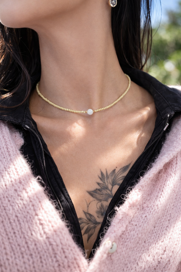 Tuvplāna lifestyle fotogrāfija saulainā dienā ar sievietes kaklu un atslēgas kaulu līniju fokusā. Ap kaklu – smalks gaiši dzeltenu pērlīšu čokeris ar centrālu baltu mēnessakmens bumbiņu. Silta saules gaisma izceļ ādas mirdzumu un akmens spīdumu, redzams ziedu tetovējums uz krūtīm, fons maigi izpludināts.