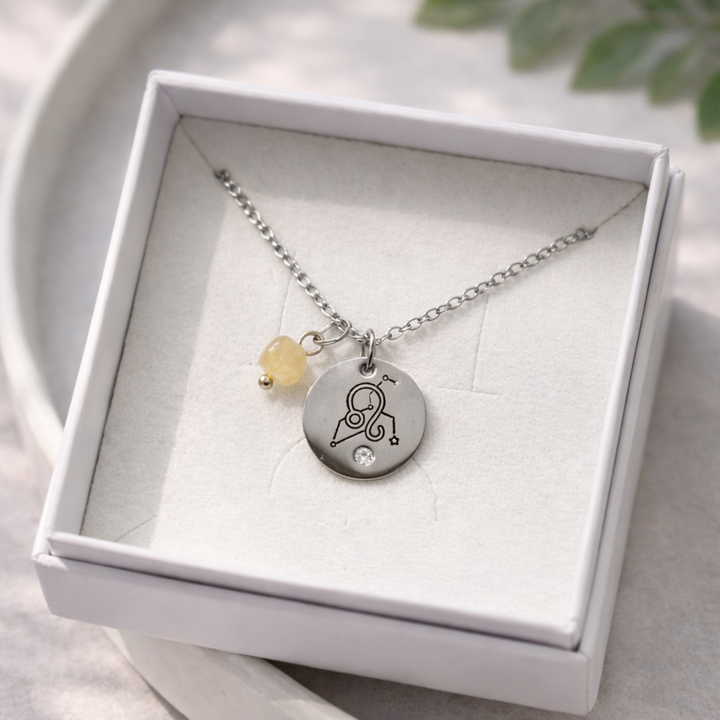 Sudraba kaklarota ar apaļu gravētu kulonu un nelielu dzeltenīgu akmens piekariņu, ievietota baltā rotaslietu kastītē uz balta keramikas šķīvja, ar maigu dabīgo apgaismojumu un izpludinātu zaļu augu fonā.
