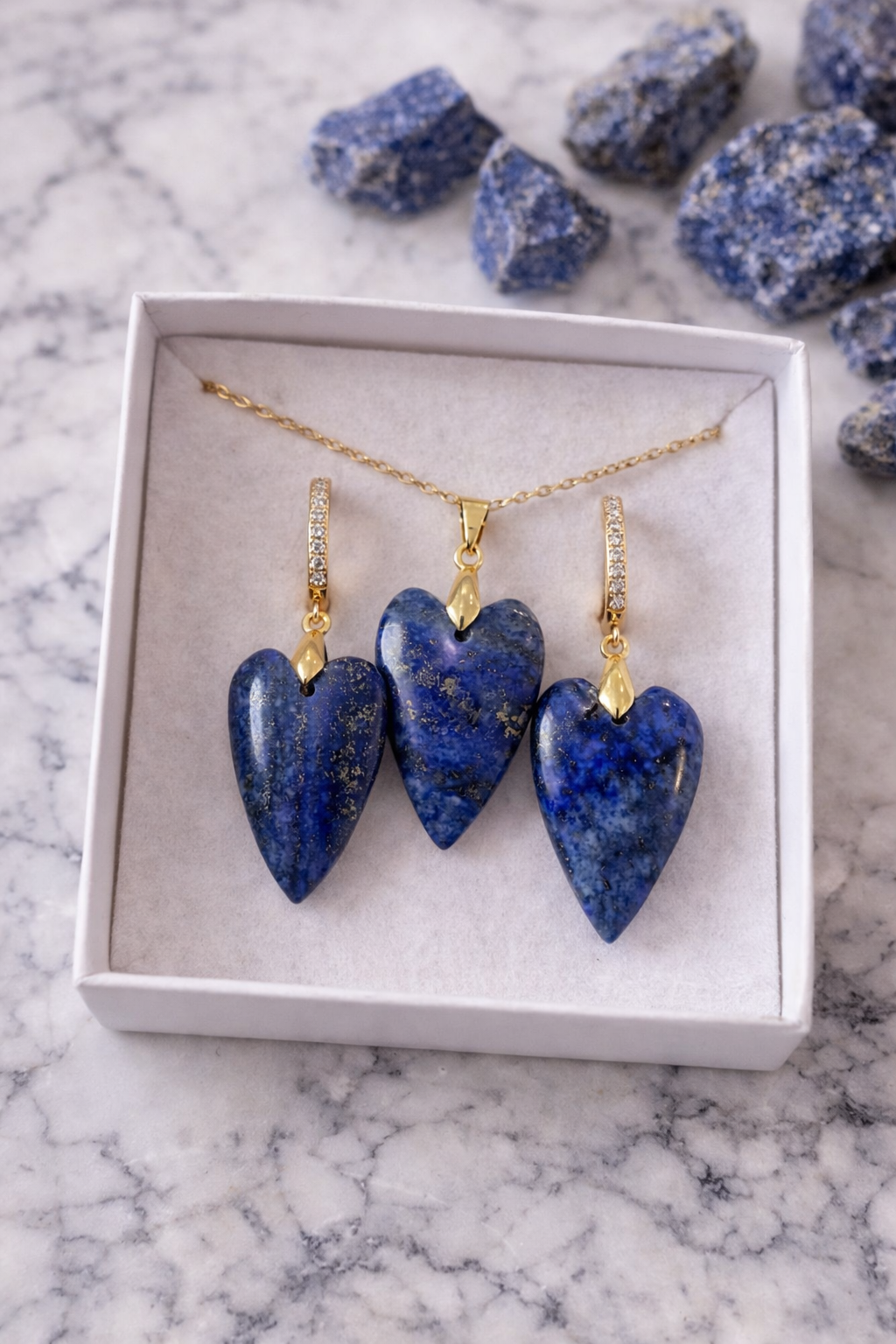 Zelta kaklarota un auskari ar lazurīta sirds formas kuloniem, eleganti izkārtoti baltā kastītē uz marmora fona, fonā redzami neapstrādāti lazurīta akmens gabali.