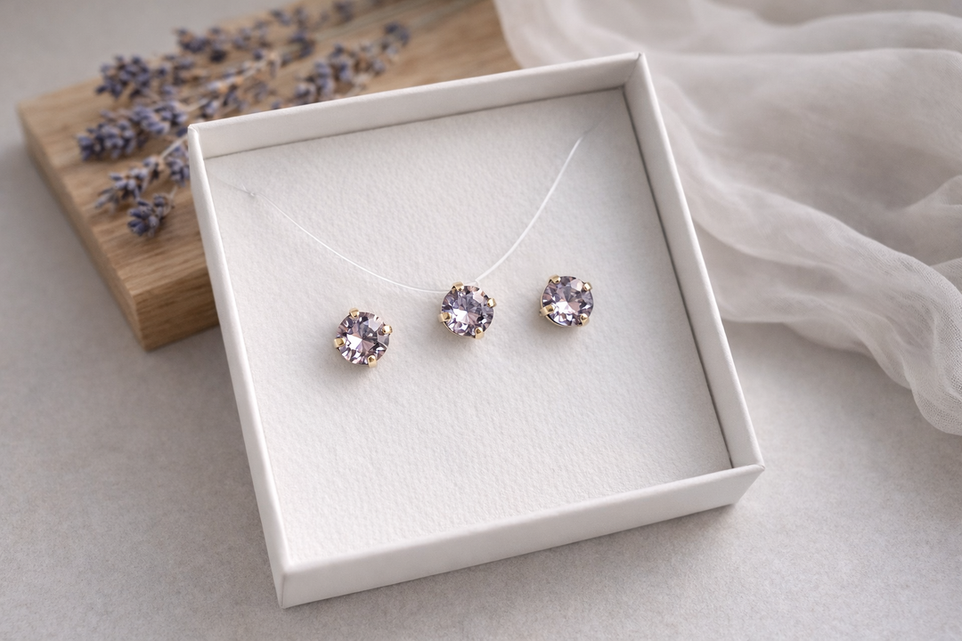 Minimālistiska rotu komplekta produktu fotogrāfija baltā kastītē – sudraba ietvarā iestrādāti cerinu krāsas kristāli.Komplektā nagliņu auskari un kaklarota uz caurspīdīgas auklas. Fons gaišs, ar koka dēlīti, žāvētu lavandu un viegli izpludinātu audumu, maigs dabīgais apgaismojums.