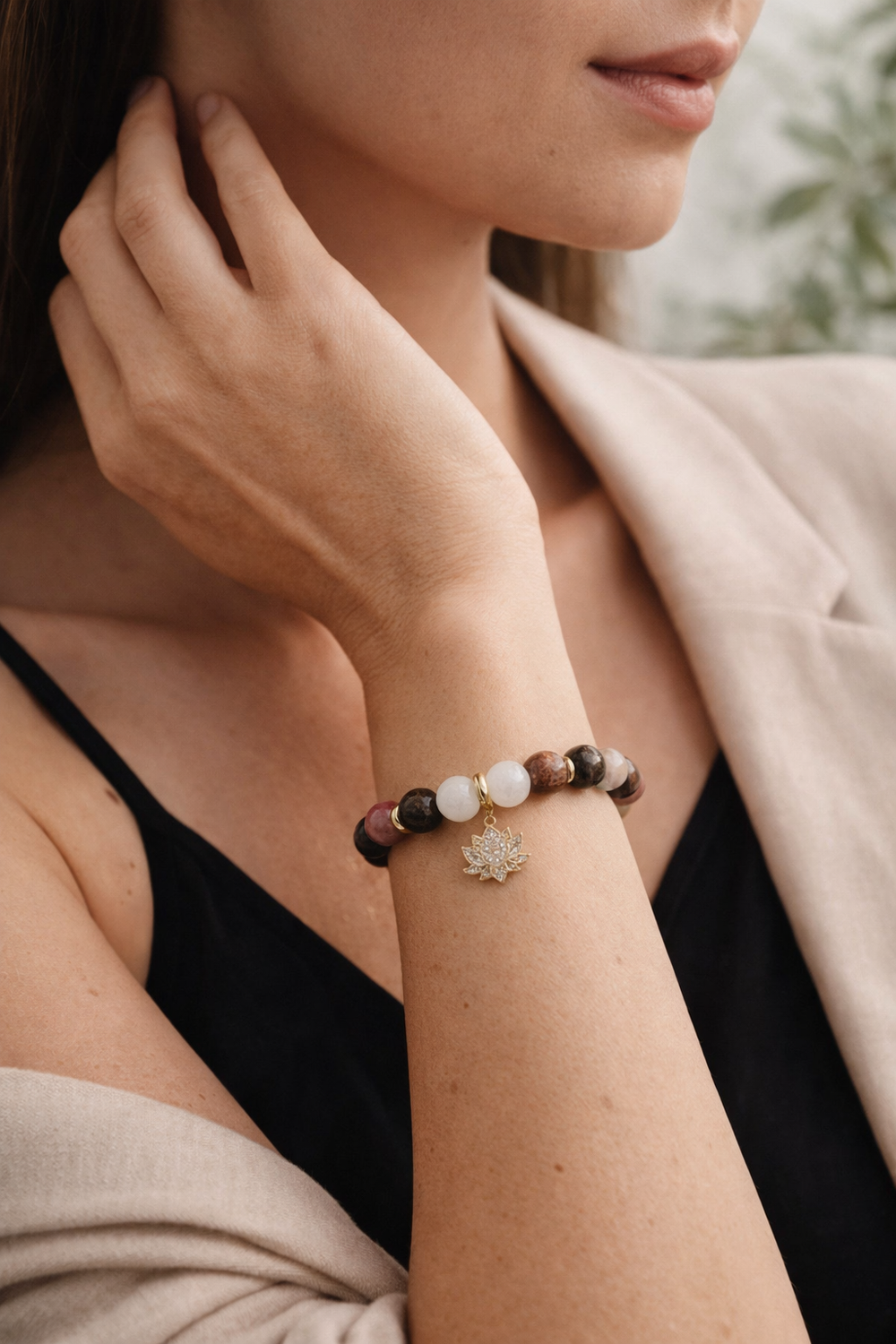 Redakcionāla tuvplāna fotogrāfija ar sievietes roku pie kakla, valkājot dabīgo akmeņu aproci ar mēnessakmeni, pirītu un rodonītu un zelta lotosa piekariņu; sievietei melns tops ar plānām lencītēm un gaiša bēša žakete, fons maigi izpludināts ar zaļu augu elementu.