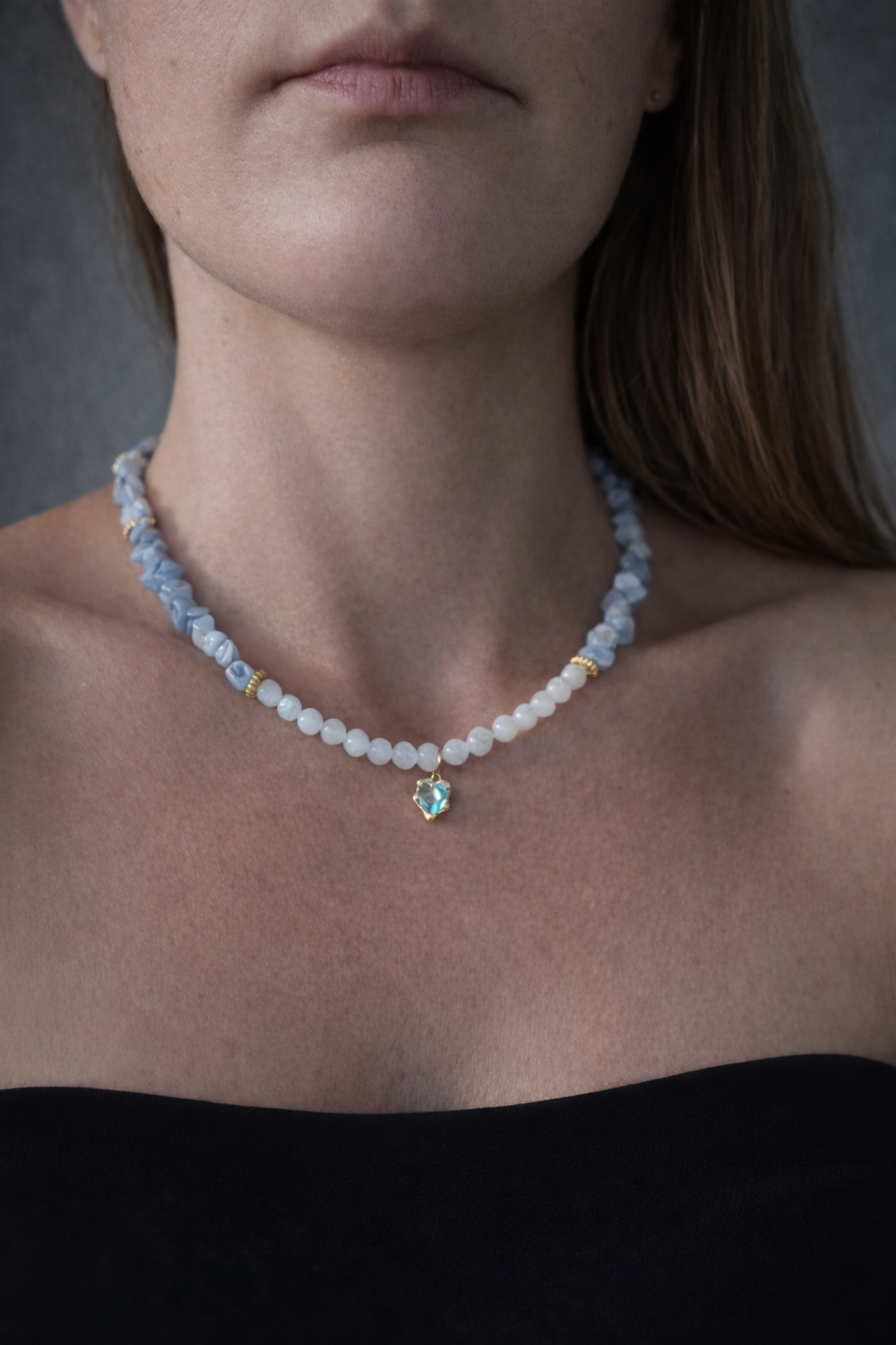 Tuvplāna portrets ar sievietes kaklu un atslēgas kauliem, valkājot kaklarotu no gaiši ziliem akmens čipsiem un baltām pērlītēm ar zeltītu sirds formas kulonu; tumšs tops un neitrāls, maigi izpludināts fons.
