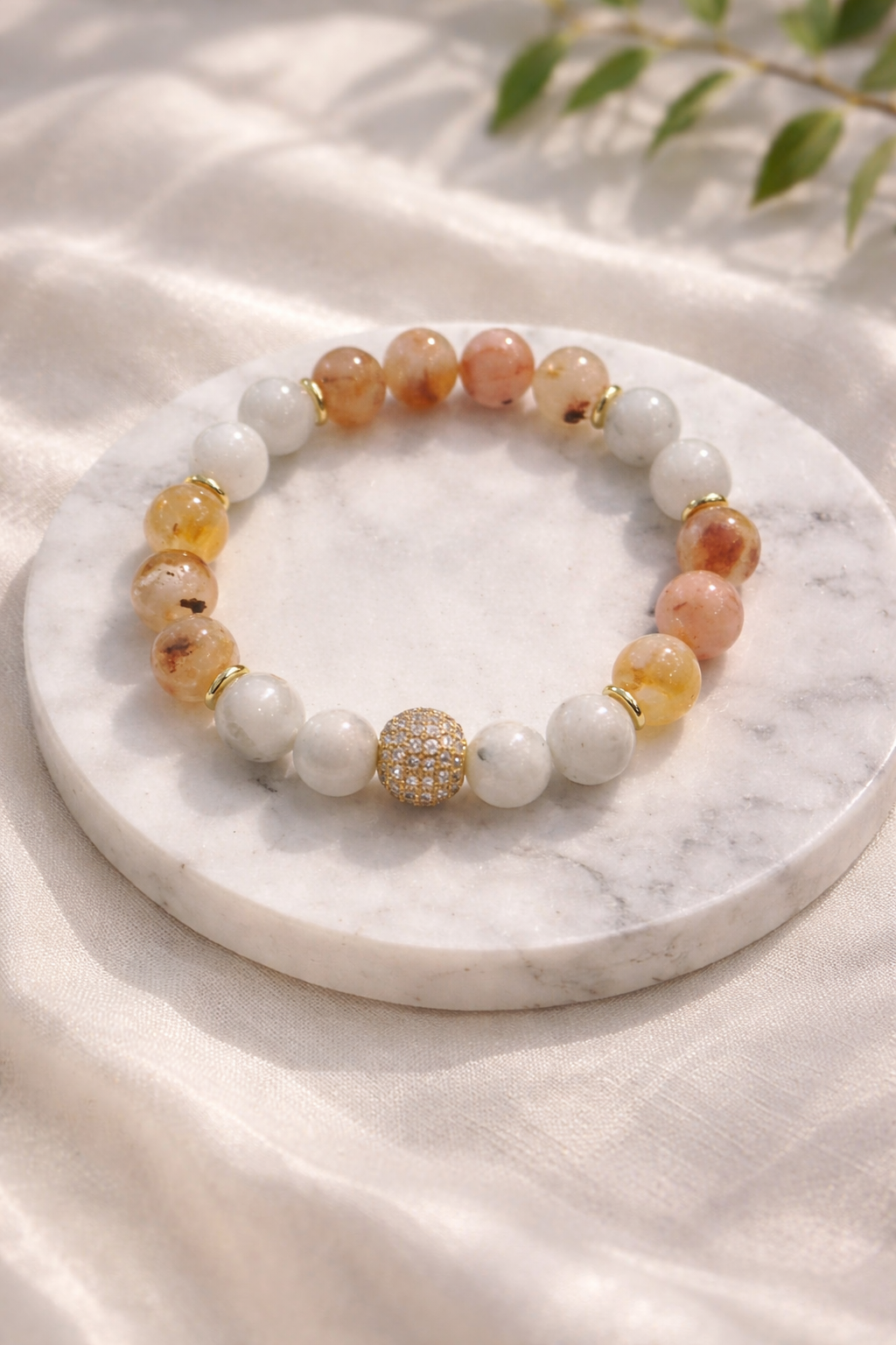 Dabīgo akmeņu aproce ar mēnessakmens un citrīna pērlēm un zelta akcenta lodīti, novietota uz balta marmora paliktņa ar gaiša zīda fonu un maigi izpludinātu zaļu augu zaru fonā.