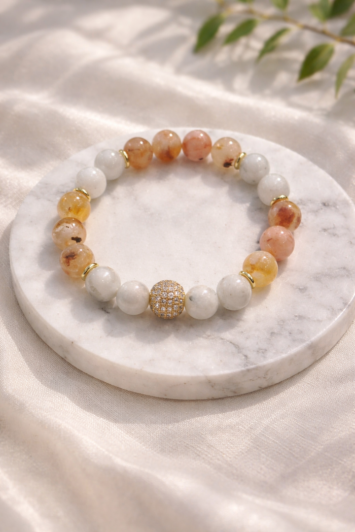 Dabīgo akmeņu aproce ar mēnessakmens un citrīna pērlēm un zelta akcenta lodīti, novietota uz balta marmora paliktņa ar gaiša zīda fonu un maigi izpludinātu zaļu augu zaru fonā.