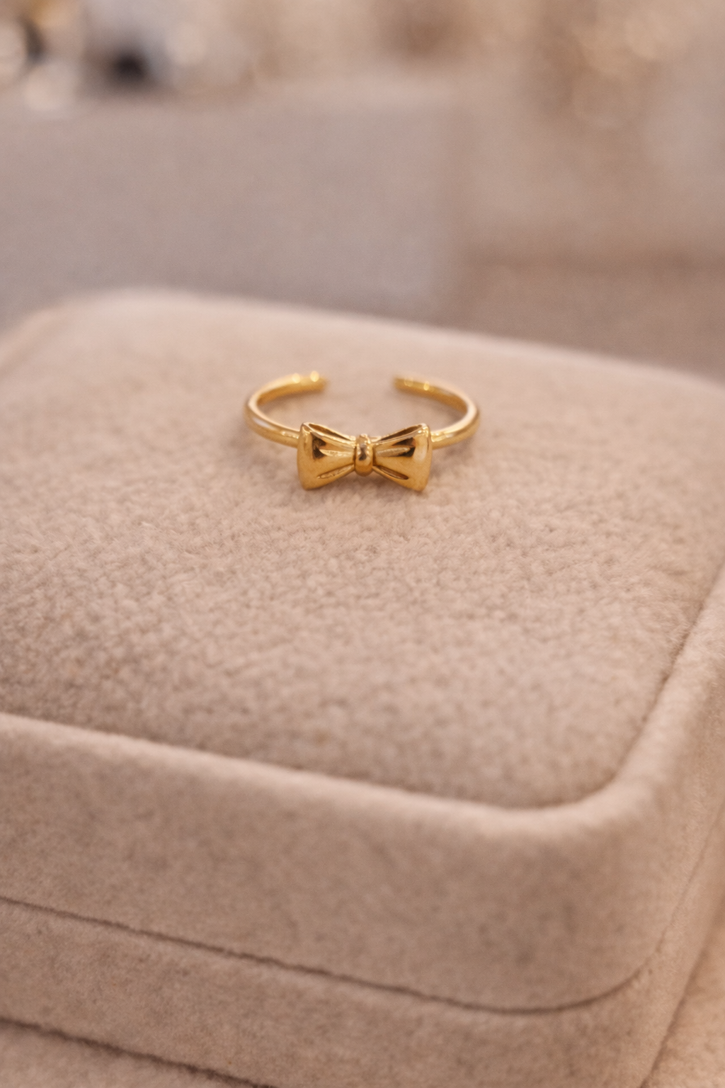 Smalks zelta atvērta dizaina gredzens ar nelielu bantītes motīvu centrā, novietots uz gaišas samta juvelierizstrādājumu kastītes maigā, siltā apgaismojumā.